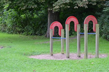 Ahşap direkler üzerinde üç büyük, kırmızı, at nalı şekilli oyun alanı ekipmanı, arka planda ağaçlar olan çimenli bir parkta, Flensburg Ostseebad 'da antrenman yapmak, yukarı çekmek ve pratik yapmak için kullanılır.