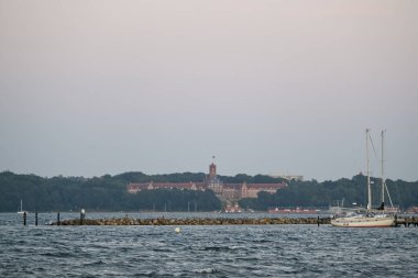 Marinefernmeldeschule Flensburg artık ağaçlar ve suyla çevrili stratejik keşif okuludur. Bir yelkenli, sakin dalgalarla ön planda demirler..
