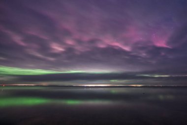 Sakin bir su kütlesinin bulunduğu sakin bir manzara bulutlu bir gecede, yeşil ve mor tonlarını da içeren canlı renklerle dolu dramatik bir gökyüzünü yansıtıyor..