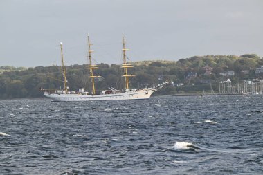Yeşil tepeler ve arka planda bir kıyı kasabasıyla çevrili üç direkli Alman Donanma Gemisi Gorch Fock sakin bir denizde yelken açıyor. Gökyüzü parçalı bulutlu, sakin bir atmosfer yaratıyor..