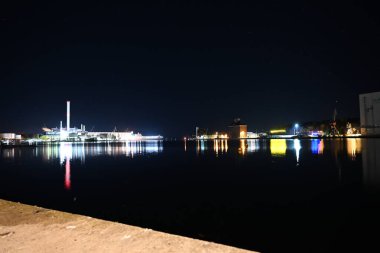 Işıklı binaları ve sudaki yansımaları olan sakin bir rıhtım manzarası. Sahnede sanayi yapıları ve sakin bir atmosfer yer alıyor..