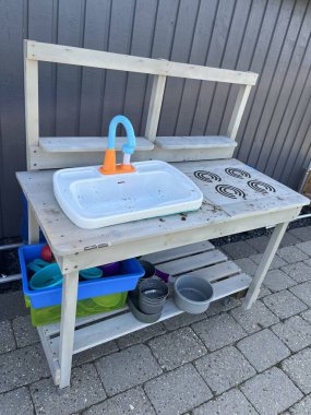 Lavabosu, ocağı ve renkli plastik tabakları olan ahşap bir masa içeren açık havada oynanan bir oyun. Masa eskimiş ve çeşitli oyun öğeleriyle çevrili asfalt bir yüzeye yerleştirilmiş..