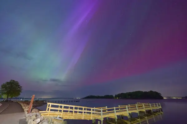 Kuzey Işıkları 'nın çarpıcı bir görüntüsü, sakin bir su kütlesinin üzerinde mor, yeşil ve mavi renkli canlı renklerle gece gökyüzünü aydınlatır. Arka planda kayalık kıyılar ve ağaçlarla çevrili ahşap bir rıhtım suya doğru uzanır..