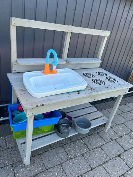 Lavabosu, ocağı ve renkli plastik tabakları olan ahşap bir masa içeren açık havada oynanan bir oyun. Masa eskimiş ve çeşitli oyun öğeleriyle çevrili asfalt bir yüzeye yerleştirilmiş..