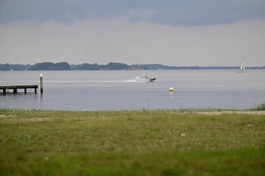 Suyun üzerinde hızla giden bir teknenin olduğu sakin bir göl manzarası, uzak bir yelkenli ve ön planda ahşap bir rıhtımla. Gökyüzü bulutlu, sakin bir ortam ekliyor..