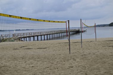 Arka planda suya uzanan ahşap iskeleli iki voleybol ağının yer aldığı kumlu bir plaj sahnesi. Gökyüzü bulutlu, sakin bir atmosfer yaratıyor..