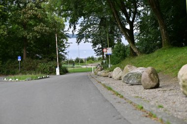 Yeşil park alanına giden sessiz bir yol. Bir tarafında büyük taşlar var. Ağaçlar gölge sağlar ve arka planda bir işaret direği görünür. Huzurlu ve doğal bir sahne..