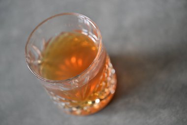 Kehribar rengi sıvıyla dolu kristal bir bardağın yakından görüntüsü, desenli gri bir yüzeye yerleştirilmiş. Cam karmaşık tasarımlar içeriyor, ışığı güzel yansıtıyor..