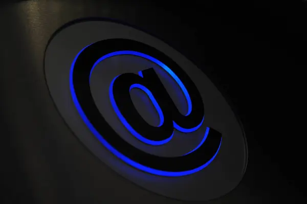 Metalik bir yüzeyde parlayan mavi @ sembolünün yakın çekimi, modern ve teknolojik bir estetik yaratıyor.