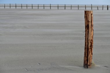 Issız bir ahşap direk, kısmen ince kumla kaplı, kumlu bir arazide duruyor. Arka planda, bir dizi çit direği görünür, derinlik ve perspektif hissi yaratır.