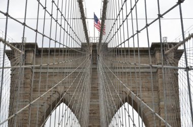 Brooklyn Köprüsü 'ndeki taş kulelerin ve süspansiyon kablolarının yakın görüntüsü. Üzerinde bulutlu bir gökyüzüne karşı dalgalanan bir Amerikan bayrağı var..