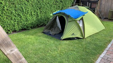 Çimenli bir alana kurulmuş yeşil bir kamp çadırı. Üzerinde mavi bir branda var. Çadır kısmen açık, içini gösteriyor. Çevredeki yeşillik ve ahşap yapı arka planda görülebilir..