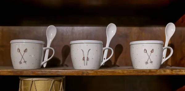 Üç dekoratif seramik kupa, kapaklı ve kaşıklı, bir kayak tasarımı, ahşap bir rafta dizilmiş. Bardaklar ince desenli beyaz ve rahat bir ortama yerleştiriliyor..