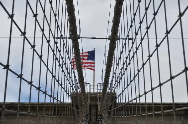 Arka planda bulutlu bir gökyüzünün altında Amerikan bayrağına giden kablolu bir asma köprü manzarası..
