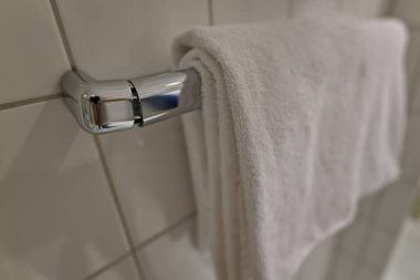 Banyodaki krom havlu askısına asılı kırık beyaz bir havlunun yakın plan fotoğrafı. Arka planda fayanslı duvarlar var..
