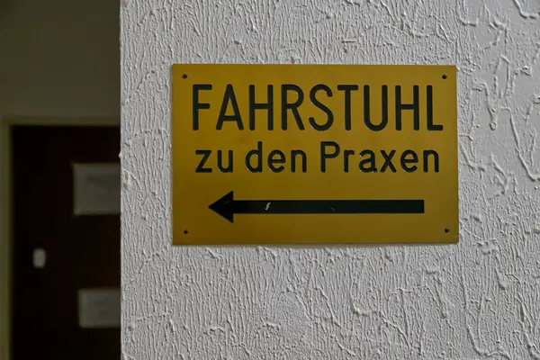 Almanca siyah metinli sarı bir tabela. Tıbbi uygulamalar için asansörün yönünü gösteriyor..