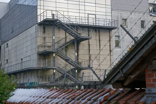 Diğer çağdaş yapılarla çevrili, metalik yangın merdiveni olan modern bir bina. Önplanda, karlı kırsal bir çatı kentsel ortama kontrastlık katar.