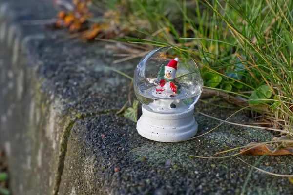 İçinde bir kardan adam olan kırık bir kar küresi. Etrafı çimlerle çevrili beton bir yüzeye yerleştirilmiş.,