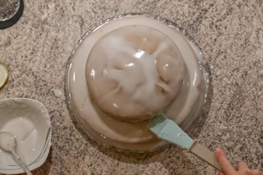 Yuvarlak, soğuk bir tatlı granit tezgahta hazırlanıyor. Bir el açık mavi bir çatalla yüzeyi fırçalarken, küçük bir kaşık yanında. Tatlının pürüzsüz, parlak bir sonu var ve etrafında köpük tabakası var..