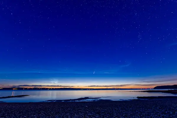 Sakin bir denizin üzerindeki sakin bir alacakaranlık sahnesi yıldızlı bir gökyüzü ve görünen bir kuyruklu yıldızın ipucuyla. Ufuk, gece çöktüğünde sıcak renklerle parlar, suyu yansıtır..