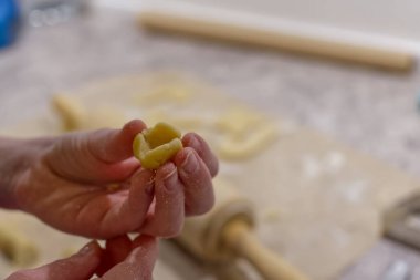 Makarna hamurunu elle şekillendiren küçük bir parça makarna sergileyen biri. Arka planda merdane ve un kaplı bir yüzey var. Bu da yemek pişirme ortamını gösteriyor..