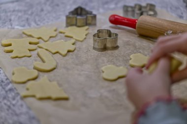 Kurabiye hamuru yapıp şekiller yaratmak için kurabiye keskisi kullanan bir çocuk. Ağaçlar, yıldızlar ve kalpler gibi çeşitli kurabiye şekilleri, tezgahın üzerinde merdane ve metal kesicilerle birlikte görülebilir.