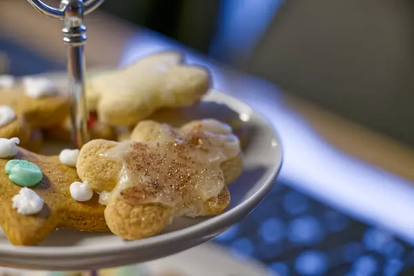Süslü zencefilli kurabiyelerden oluşan, çeşitli şekillerde ve kremalı bir tabağın yakın çekimi. Kurabiyeler klavyenin bulanık arka planına sahip katmanlı bir stantta düzenlenir..