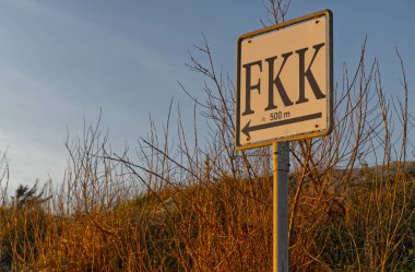 'FKK' ve 500 metre uzaklıktaki çıplaklar plajının yönünü gösteren bir işaret. Arkaplanda gün batımında kuru çimenler ve açık bir gökyüzü yer almaktadır..
