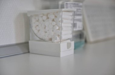Beyaz pamuklu rulo ile dolu şeffaf bir kap temiz, beyaz bir yüzey üzerinde oturur. Arka planda, hastane, klinik veya dişçi muayenehanesini anımsatan bulanık tıbbi malzemeler görülebilir