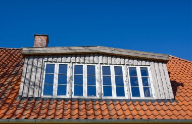 Açık mavi gökyüzünün altında birden fazla penceresi olan ahşap bir çatı yapısının yakın görüntüsü. Çatıda, doğal malzemeler ve mimari tasarım karışımı sergilenen kırmızı fayanslar ve bir baca bulunuyor..