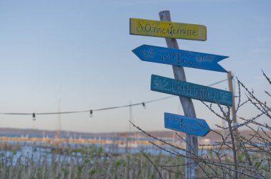 Wassersleben Flensburg 'da renkli ahşap bir tabela, oklar güneş terasını işaret ediyor, dondurma, büfe, bisiklet, arka planda mavi gökyüzü olan güzel bir marina manzarası var.