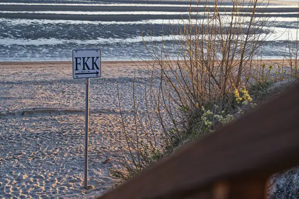 Çıplaklığı teşvik eden FKK (Freikoerperkultur) tabelasını gösteren bir plaj sahnesi. Kumlu sahil, arka planda hafif dalgalar ve yakınlarda bitki örtüsüyle görülebilir..