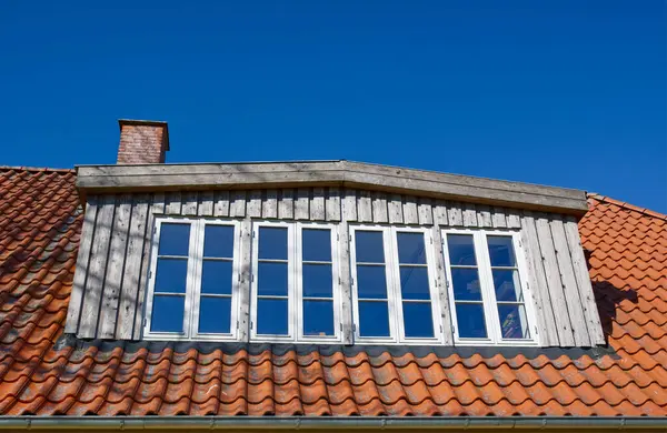 Açık mavi gökyüzünün altında birden fazla penceresi olan ahşap bir çatı yapısının yakın görüntüsü. Çatıda, doğal malzemeler ve mimari tasarım karışımı sergilenen kırmızı fayanslar ve bir baca bulunuyor..