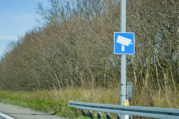 Yol kenarındaki direkte, arka planda ağaçlar ve çimenler olan mavi bir güvenlik kamerası tabelası. Sahne açık bir günde, kırsal bir otoyol ortamı sergileniyor..
