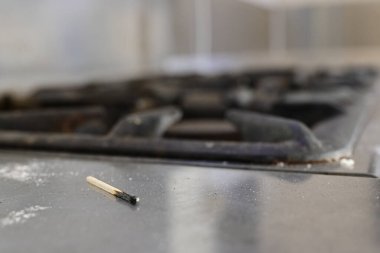 Gaz sobasının yanındaki kirli metal mutfak tezgahında yanmış bir kibritin yakın çekimi. Arka plan biraz bulanık, kibriti ve fırın ızgaralarını vurguluyor..