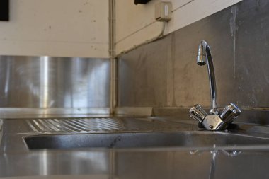 Modern musluk ve kulpları olan paslanmaz çelik bir lavabonun yakın çekim görüntüsü. Lavabo desenli bir yüzeye sahiptir ve minimalist bir tasarım sergileyen düz bir duvara dayanır..