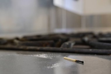 Kirli metal bir mutfak tezgahında, gaz sobasının yanında yanmış bir kibritin yakın çekimi. Arka plan biraz bulanık, kibriti ve fırın ızgaralarını vurguluyor..