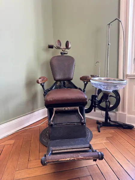 Kahverengi deri koltuklu antika bir dişçi sandalyesi, ahşap döşemeli, klasik tarzda bir odada. Metal bir dişçi tezgahı onun yanında, tarihsel ambiyansı da ekliyor..