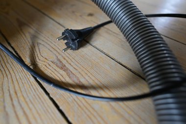 Elektrik süpürgesinin yakın plan görüntüsü elektrik kablosu ve ahşap döşeme fişi. Metalik hortum yakınlarda, modern bir ev temizliği düzeneği sergiliyor..
