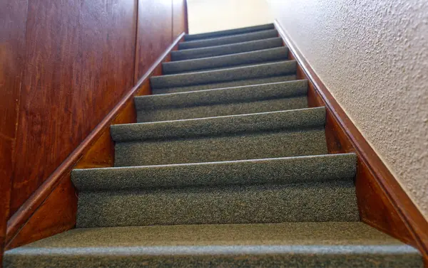 Yukarıya çıkan merdiven manzarası, halılı basamaklar ve ahşap duvarlar. Görüntü merdivenlerin altından perspektifi yakalıyor, yükselişi vurguluyor..