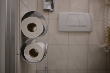 İki rulo tuvalet kağıdının modern bir tutacağa yakın çekimi. Arkaplanda, işlevsel ve temiz bir atmosfer yaratan fayanslı duvarlar ve bir telefon bulunuyor..