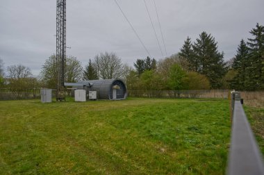 Ağaçlarla çevrili çimenli bir alanda modern bir barınağı olan bir telekomünikasyon kulesi. Gökyüzü bulutlu, sakin bir atmosfer yaratıyor..