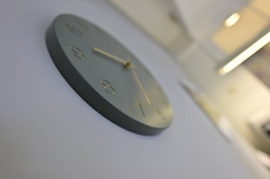 Minimalist tasarımı olan modern bir duvar saati. Gri bir yüzü, altın bir saati ve küçük elleri var. Saat açık renkli bir duvara monte edilmiş, parlak görünümünü vurguluyor..