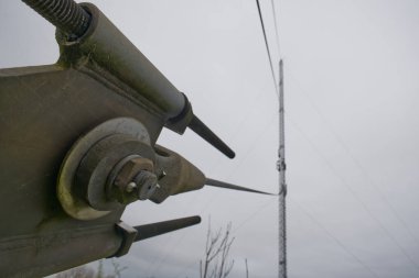 Uzun bir radyo kulesini desteklemek için kullanılan metal gerilim cihazının cıvata ve kablolara odaklı yakın çekim görüntüsü. Arka planda bulutlu bir gökyüzü ve uzaktan kule var..