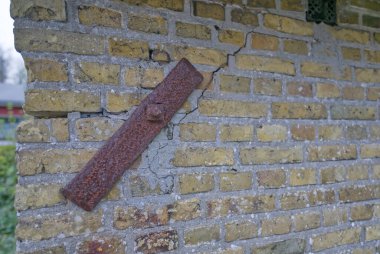 Çatlakları ve paslı metal kayışı gösteren yıpranmış tuğla bir duvar görüntüsü. Tuğlalar çeşitli sarı ve gri tonlarda ve biraz harç eksik. Sahne, yaşlılık ve çürüme hissi veriyor..
