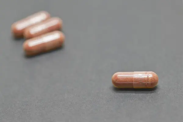 Koyu bir zemin üzerinde antioksidan ve Astaxanthin içeren kahverengi kapsüllere yakın plan, bir kapsül gruptan uzakta duruyor. Kapsüllerin parlak bir yüzeyi var ve düzenlenmişler..