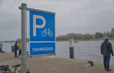 Kappeln 'de bir nehrin kenarında mavi bir bisiklet parkı tabelası, arka planda insanlar var. İşaret, bisiklet ikonunun yer aldığı, bisikletler için belirlenmiş bir alanı gösteriyor. Kappeln 'de hava bulutlu ve sakin.
