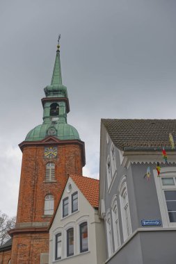 Kappeln 'deki tarihi St. Nikolai-Kirche kilisesi yeşil bakır kulesiyle bulutlu bir gökyüzüne karşı duruyor. Yakınlarda, çeşitli mimari tarzlara sahip geleneksel binalar..