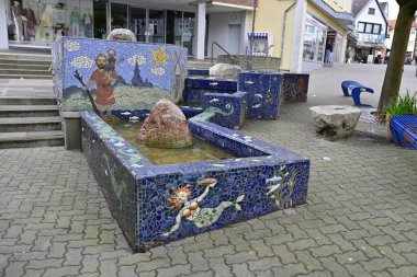 Taşlar ve asfalt bir alanla çevrili sanatsal karakter ve doğa tasarımları içeren renkli mozaik çeşmesi. Çeşme Kappeln 'de, arka planda Kappeln' de dükkanlar bulunmaktadır. .