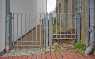 Metal bir kapı iki bina arasında kısmen açık, çimenli bir alana açılan dar bir patika var. Çevrede tuğla, taş kaldırım ve birkaç bitki var..
